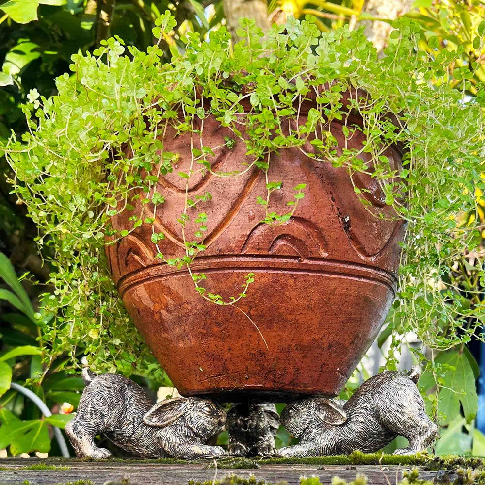 Decorative terracotta pot with greenery and three gold rabbit pot holders in a garden setting.
